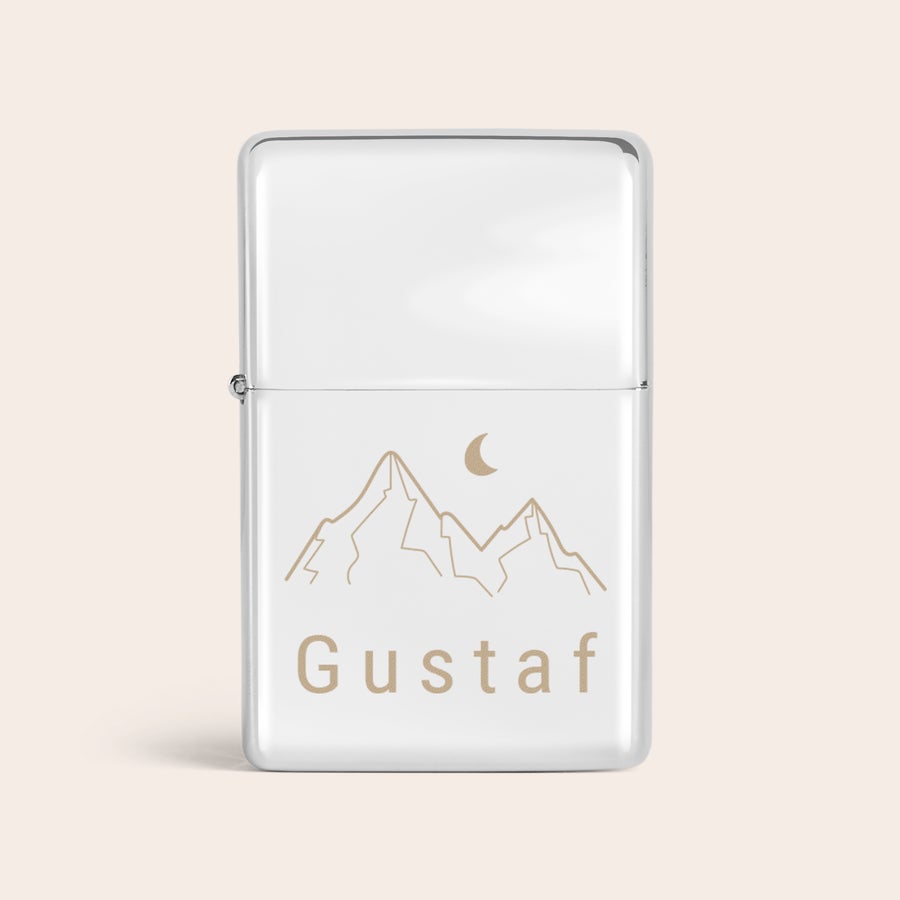 Graverade tändare En graverad namntändare i silver med bergsmotiv och mån-detalj ovanför namnet "Gustaf" graverat i guldliknande färg.