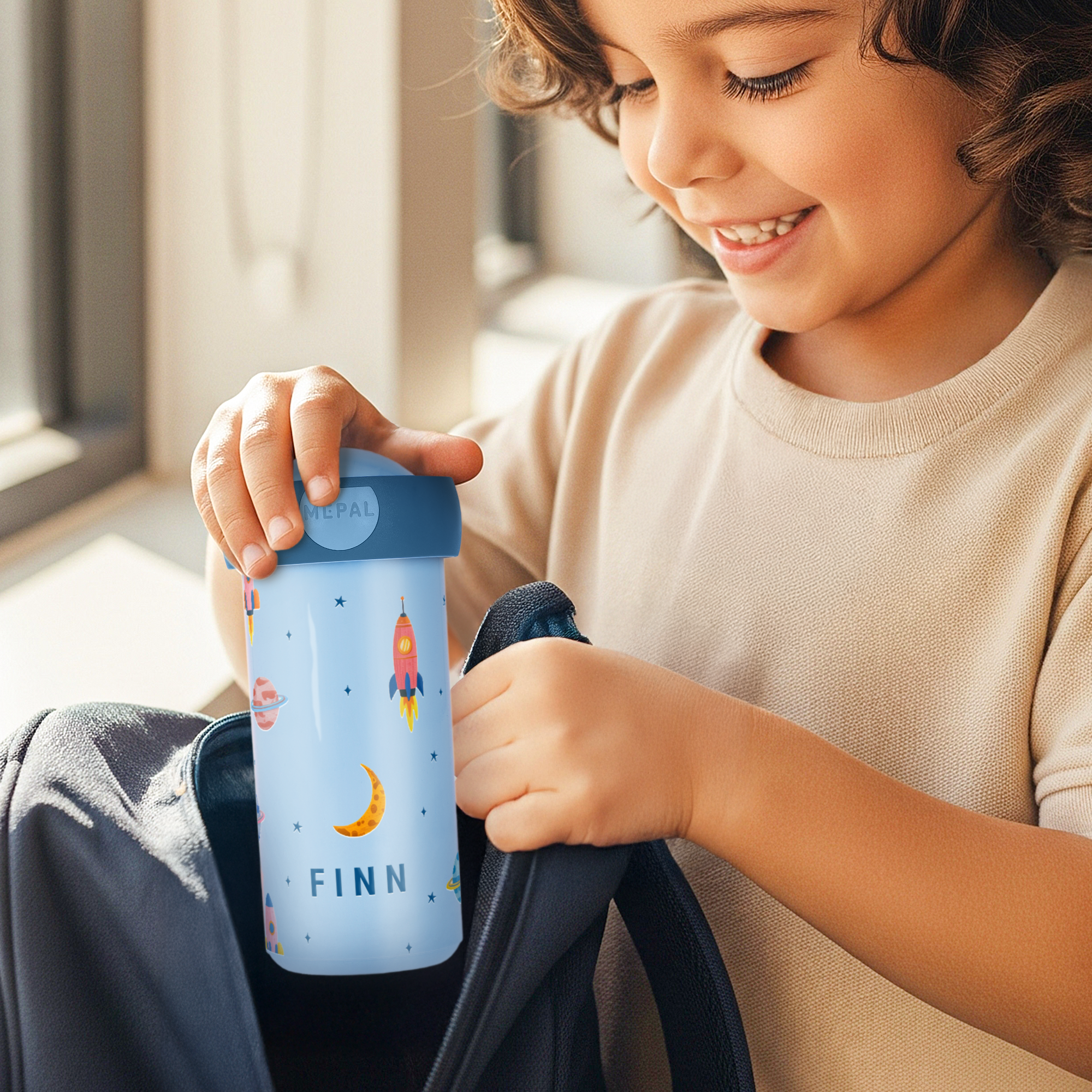 Bambino sorridente che mette una tazza scolastica personalizzata con nome Finn e grafica spaziale nello zaino. Rendi speciale ogni giorno di scuola con una tazza scolastica personalizzata.
