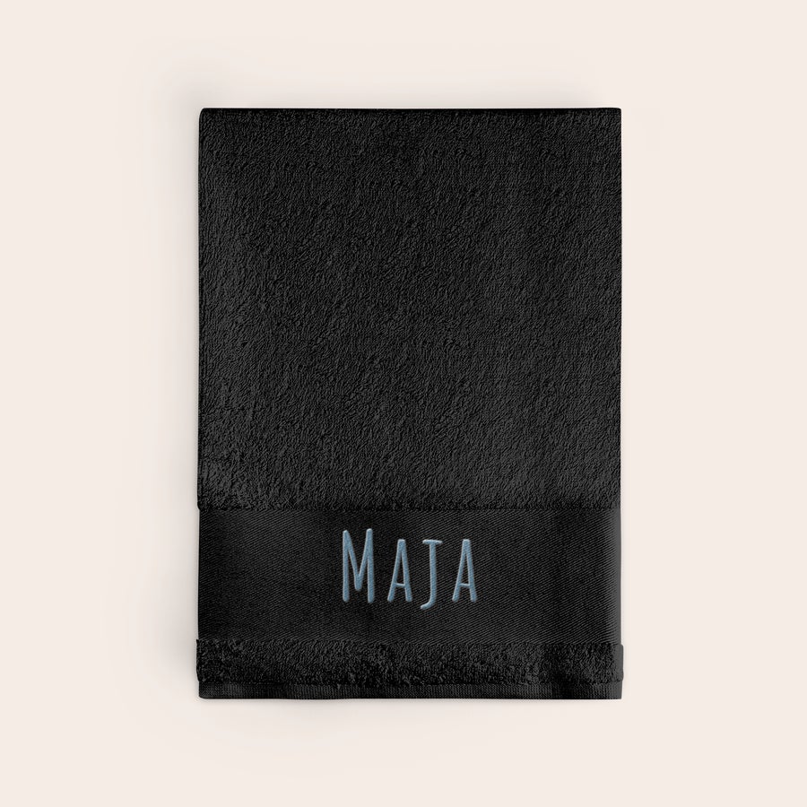 Håndkle med tekst Svart brodert håndkle med navnet MAJA i lyseblå skrift.