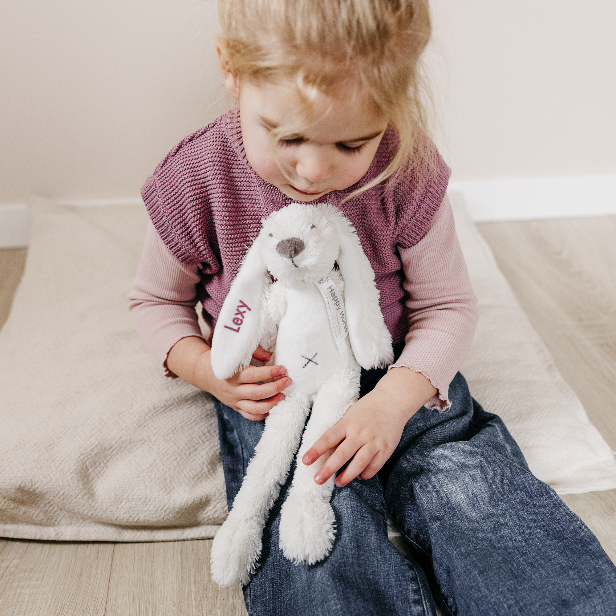 Niña rubia sentada abrazando un peluche de conejo blanco personalizado con el nombre Lexy bordado.