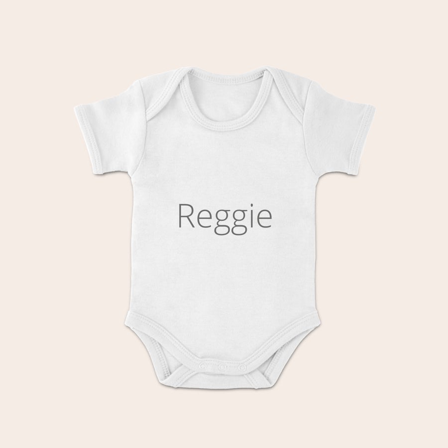 Personalizowane śpioszki niemowlęce Białe body niemowlęce z nadrukowanym imieniem Reggie na środku, zapinane na zatrzaski.