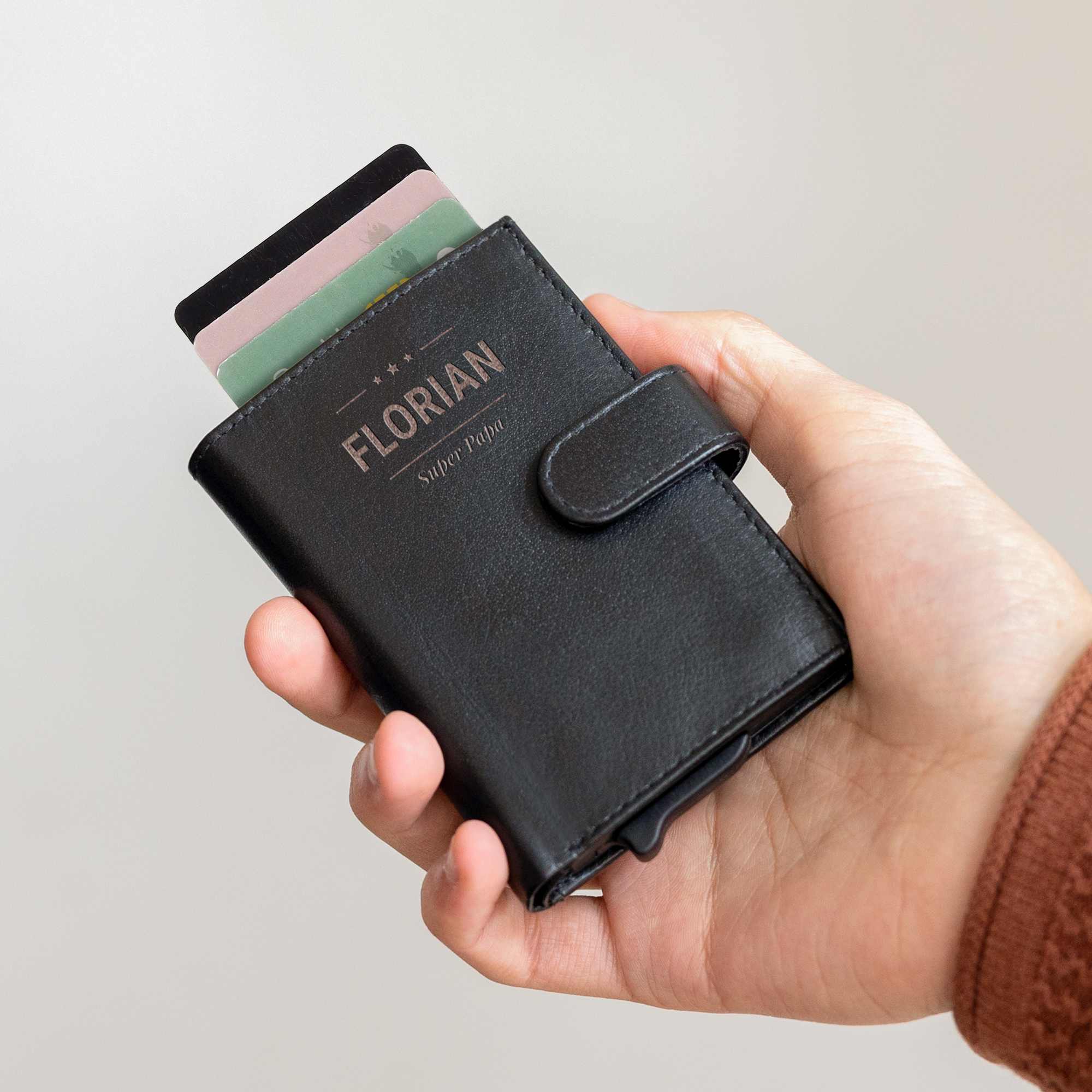 Schwarzes personalisiertes Kartenetui aus Leder in einer Hand, graviert mit dem Namen FLORIAN und "Super Papa", mit drei Kreditkarten sichtbar. Keine schweren Brieftaschen mehr mit einem eigenen Kartenetui mit Namen.