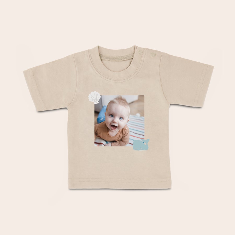 Baby T-skjorte Beige baby-t-skjorte med trykt bilde av en smilende baby, havskjell og hval for en stilig baby