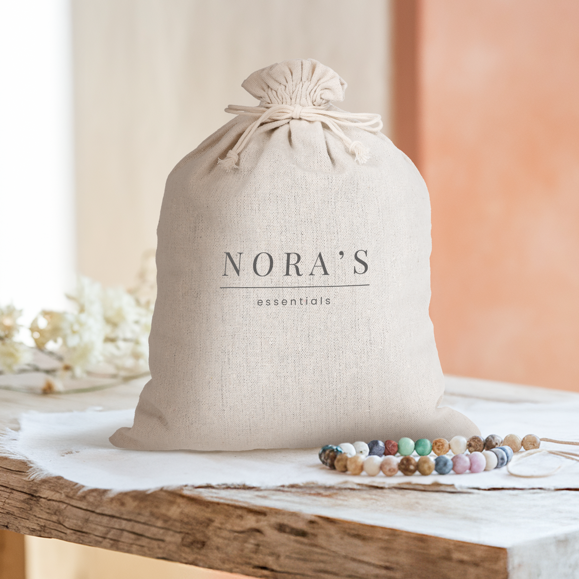 Personlig snorpose i bomull, trykket med teksten "NORA'S essentials" i svart, på et lyst trebord.