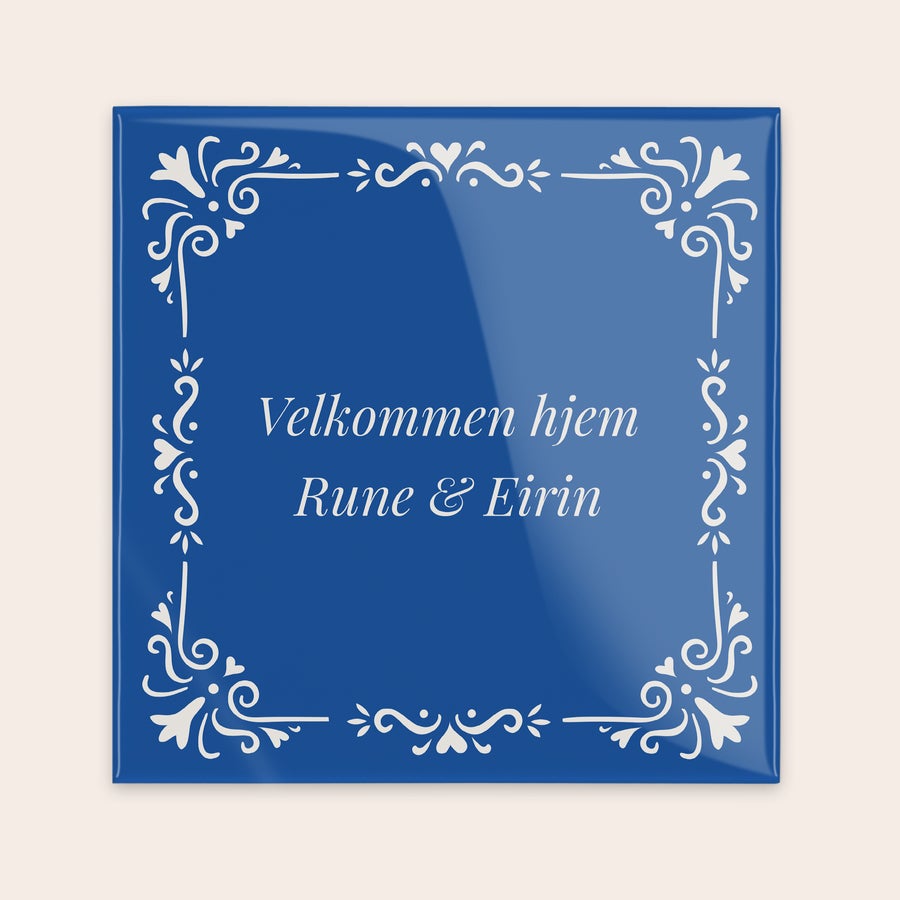 Tradisjonell flis Blå keramisk flis med hvit dekorasjon, trykt med teksten "Velkommen hjem Rune & Eirin" som et personlig ordsprog.