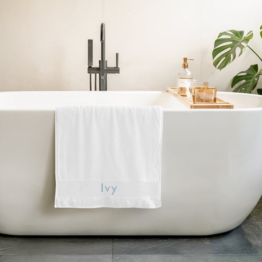 Asciugamano con testo Asciugamano super morbido e coccoloso ricamato con il nome "Ivy" in blu chiaro, appeso a una vasca da bagno bianca.