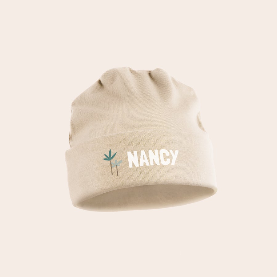 Babyhue med navn Beige personlig babyhue med navn "NANCY" og tryk af to palmer, lavet af økologisk bomuld.