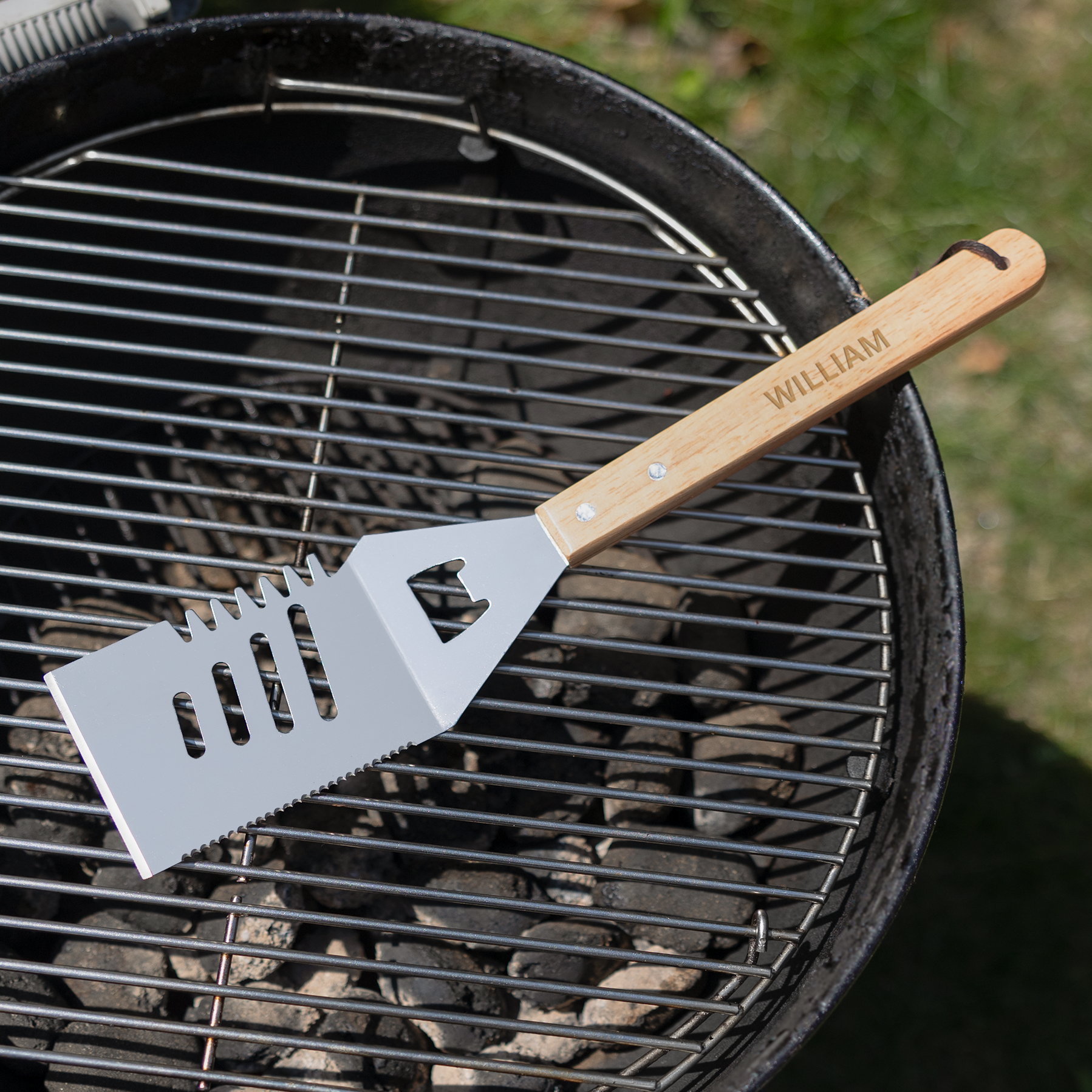 Personlig grillspade i trä och metall med namnet WILLIAM ingraverat på handtaget, ligger på grillgallret över kolbädden.