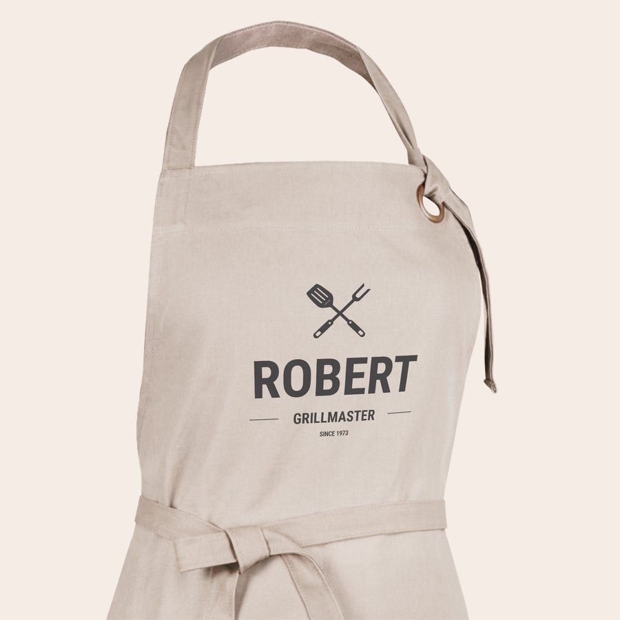 Zástera s fotkou/textom Personalizovaná zástera s menom Robert, symbolmi grilovania a textom Grillmaster Since 1973, vytlačená na béžovej látke.