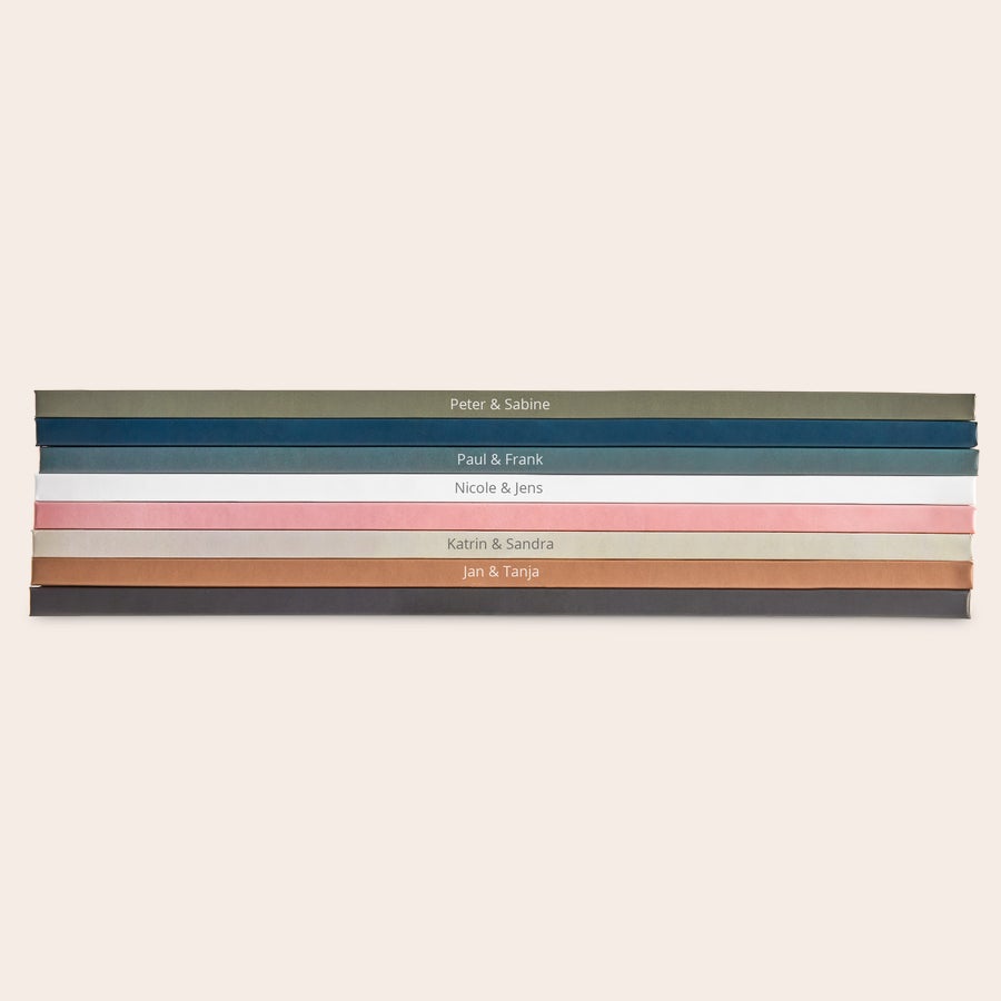 Fotobuch gestalten Stapel personalisierter Fotobücher in verschiedenen Farben mit gedruckten Namen wie Peter & Sabine, Paul & Frank zum Sammeln deiner schönsten Erinnerungen.