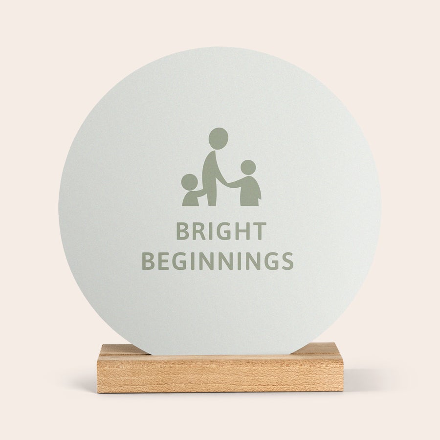 Piastrella in Legno con Foto Personalizzata Piastrella rotonda in legno con foto, personalizzata con logo "Bright Beginnings" e figure stilizzate, su base in legno.