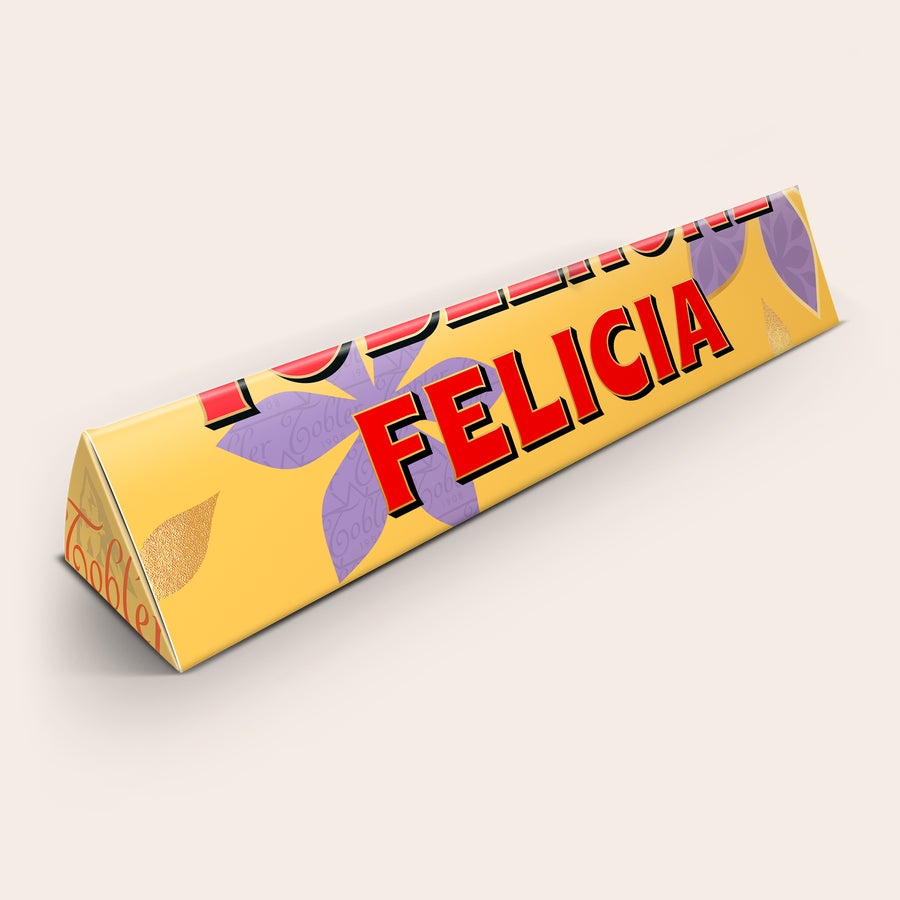 Morsdag Toblerone Personlig Toblerone-chokladkaka med namnet Felicia tryckt på det gula omslaget med lila blommor för att överraska din mamma.