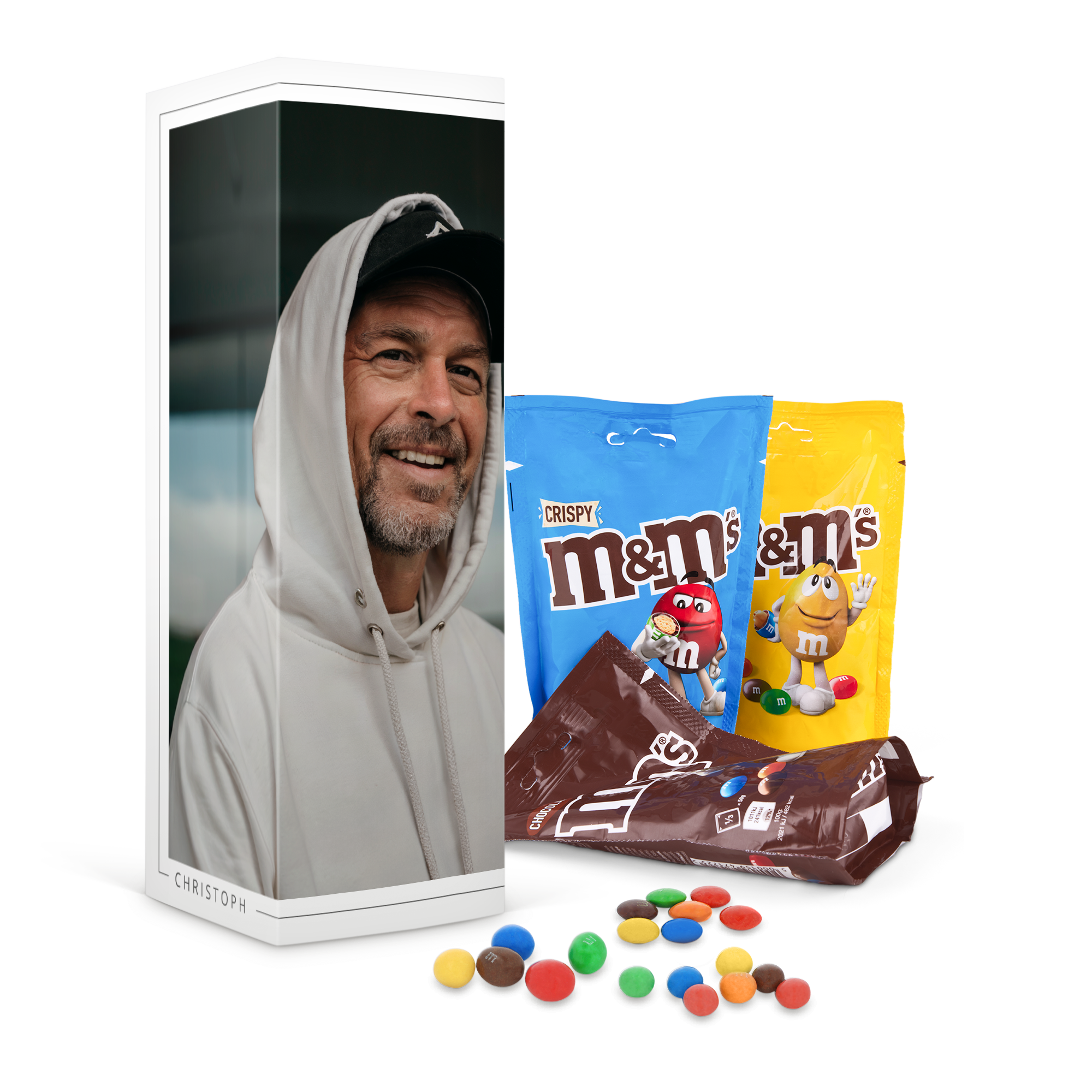 M&M's Geschenkbox bedruckt mit einem lächelnden Mann und dem Namen Christoph, umgeben von blauen, gelben und braunen M&M's Tüten und verstreuten M&M's