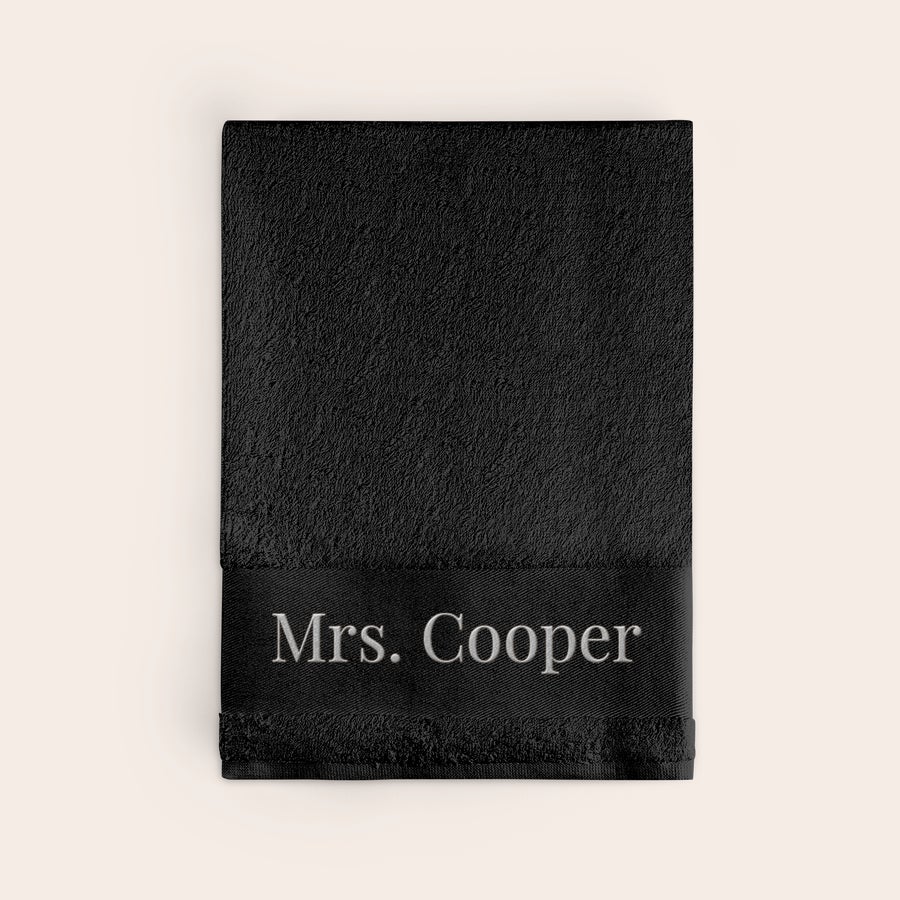 Toalha com texto Toalha preta com nome bordado "Mrs. Cooper" em branco. Torna o teu banho ainda mais pessoal com uma toalha preta bordada.