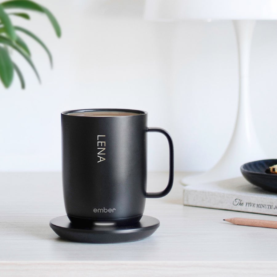 Ember Smart Control Mug Caneca Ember Control personalizada preta, com o nome "Lena" gravado a laser, sobre base de carregamento. Saboreie cada gole com um copo Ember Control personalizado!