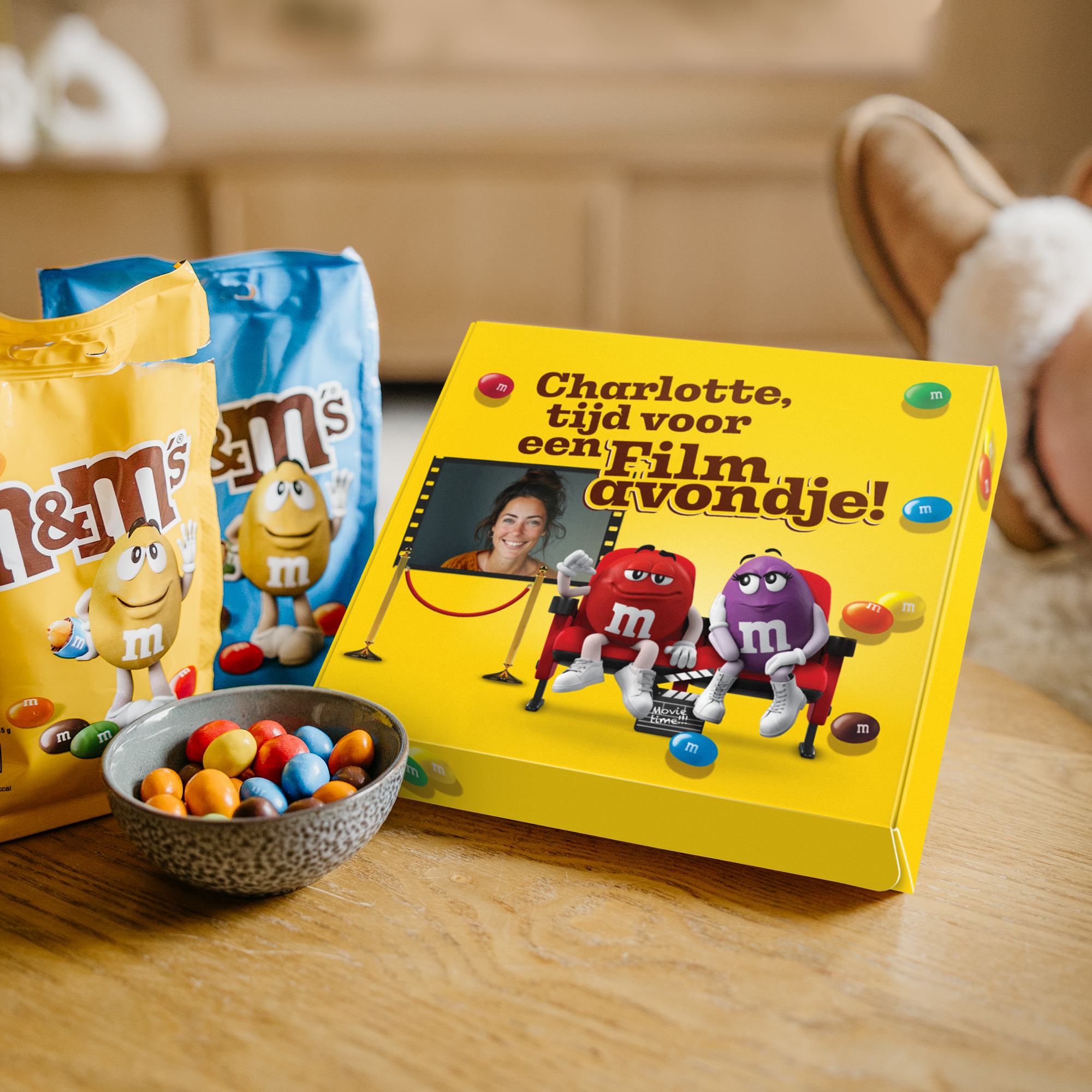 Gepersonaliseerde M&M’S Film Giftbox met de tekst 'Charlotte, tijd voor een Filmavondje!' en een foto, gedrukt op een gele doos naast twee zakken M&M’S en een kommetje snoepjes.