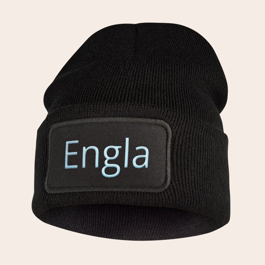 Broderad hatt Svart stickad mössa med texten "Engla" broderad i ljusblått på en svart rektangulär tyglapp.