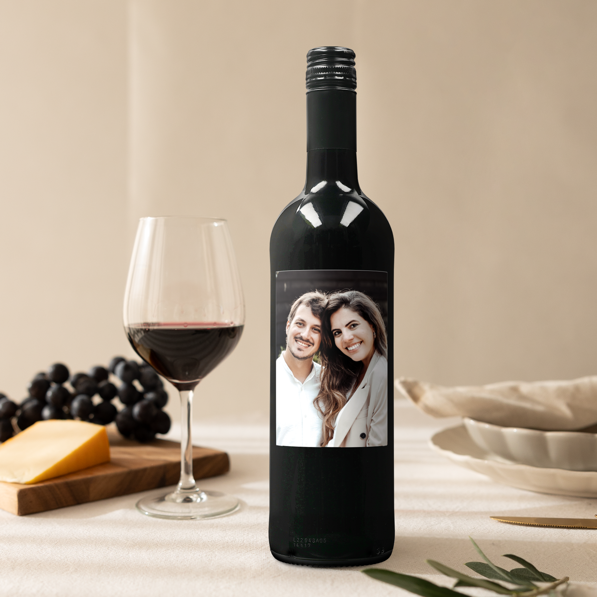 Sticlă de vin roșu Belvy cu etichetă personalizată imprimată cu fotografia unui cuplu, lângă un pahar de vin, struguri și brânză.