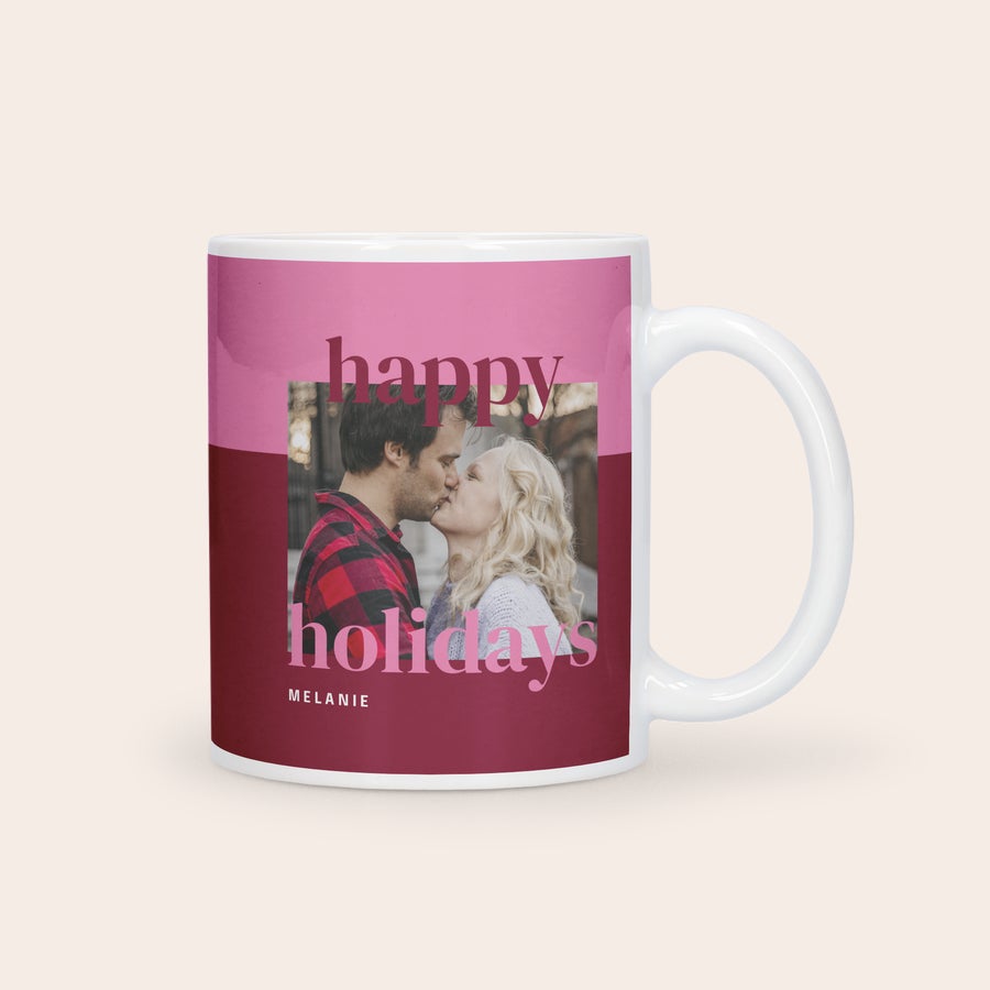 Tazza Natalizia Personalizzata con Foto Tazza bianca personalizzata con la foto di una coppia stampata e la scritta Happy holidays e il nome Melanie su sfondo rosa e rosso.