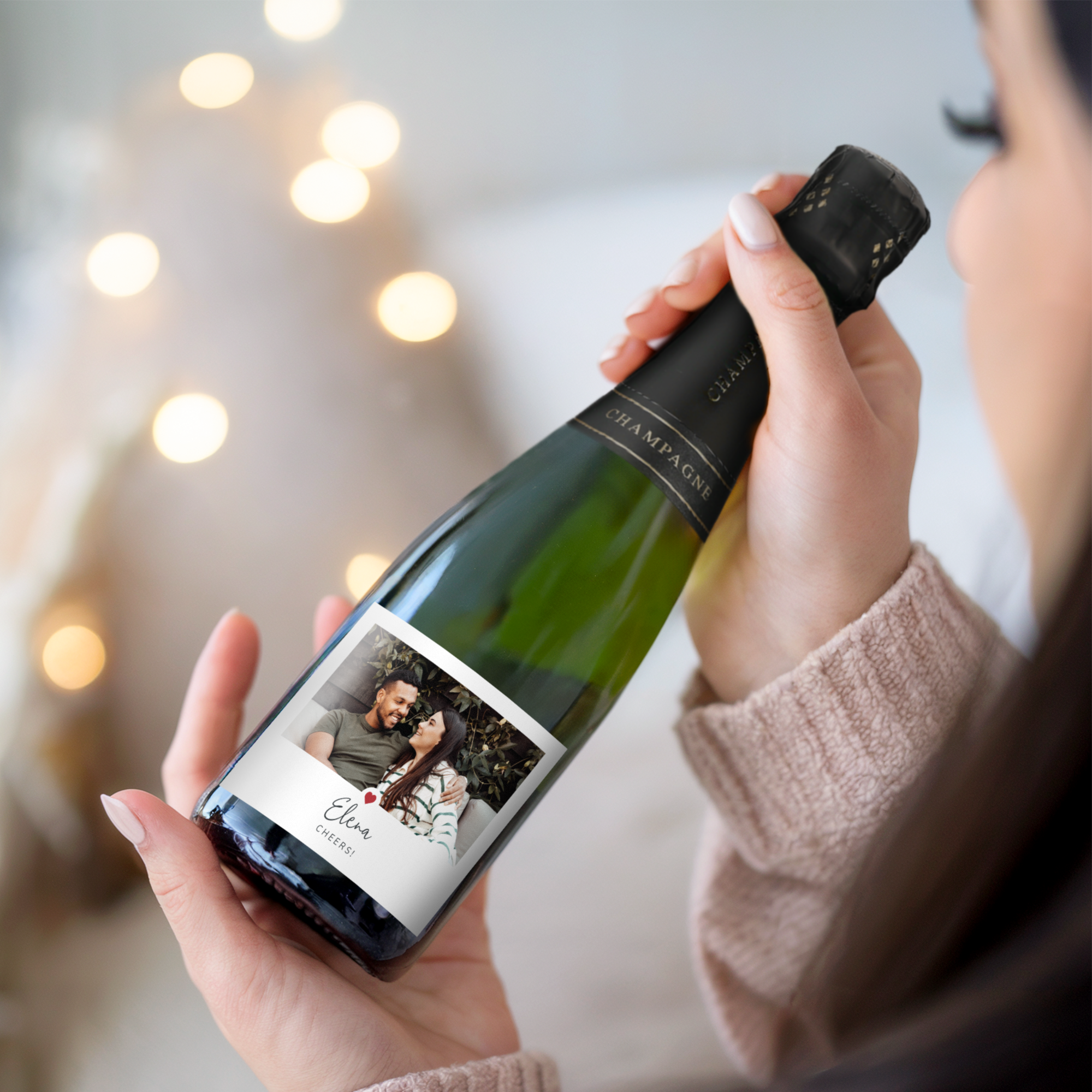 A woman holding a bottle of champagne with personalised label, printed with a photo of a couple, "Elena" and "Cheers!" text.