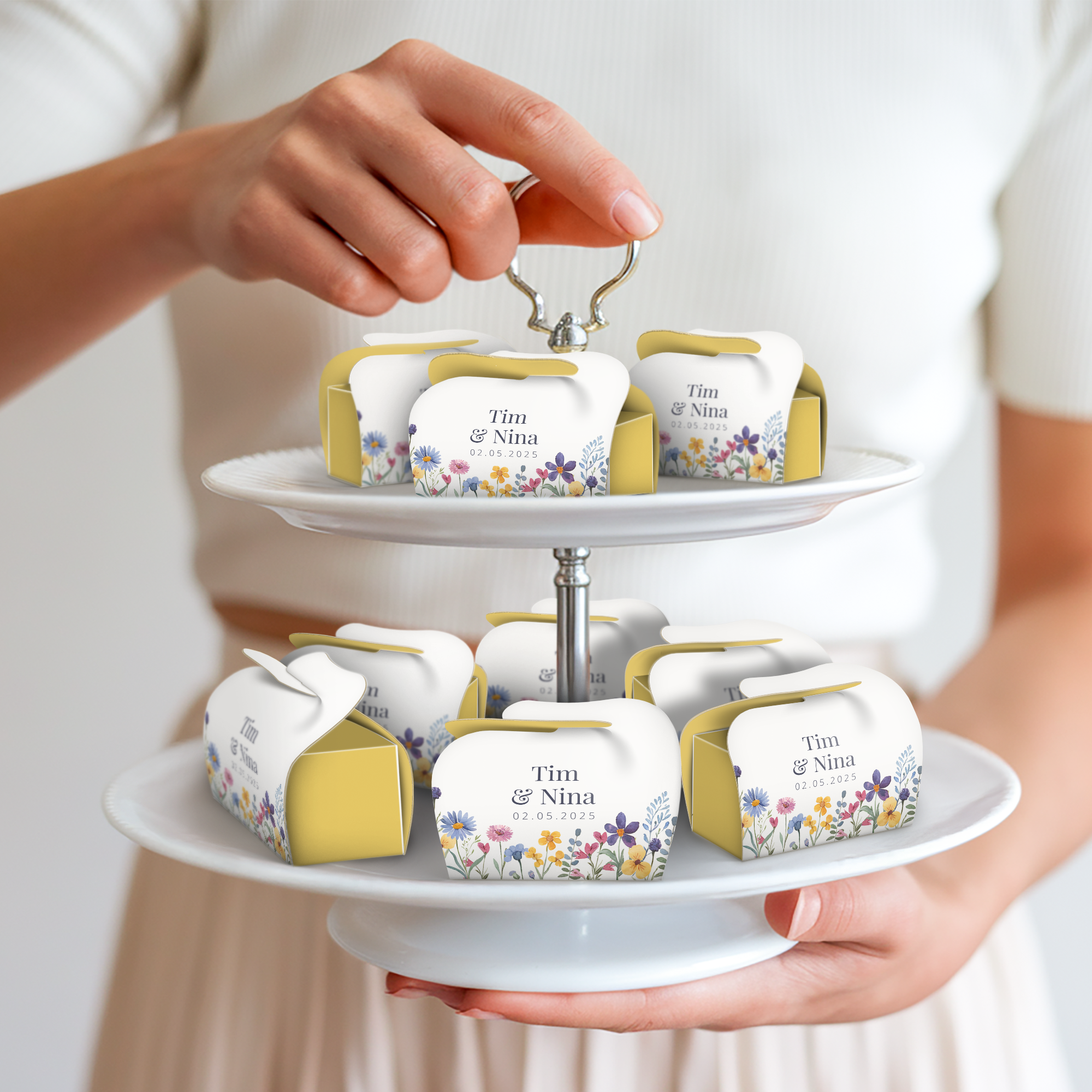 Een hand houdt een etagère vast met gepersonaliseerde Milka Favourites doosjes, bedrukt met de namen Tim & Nina en een bloemenpatroon. Ontwerp jouw zoete momenten met Milka Favourites.
