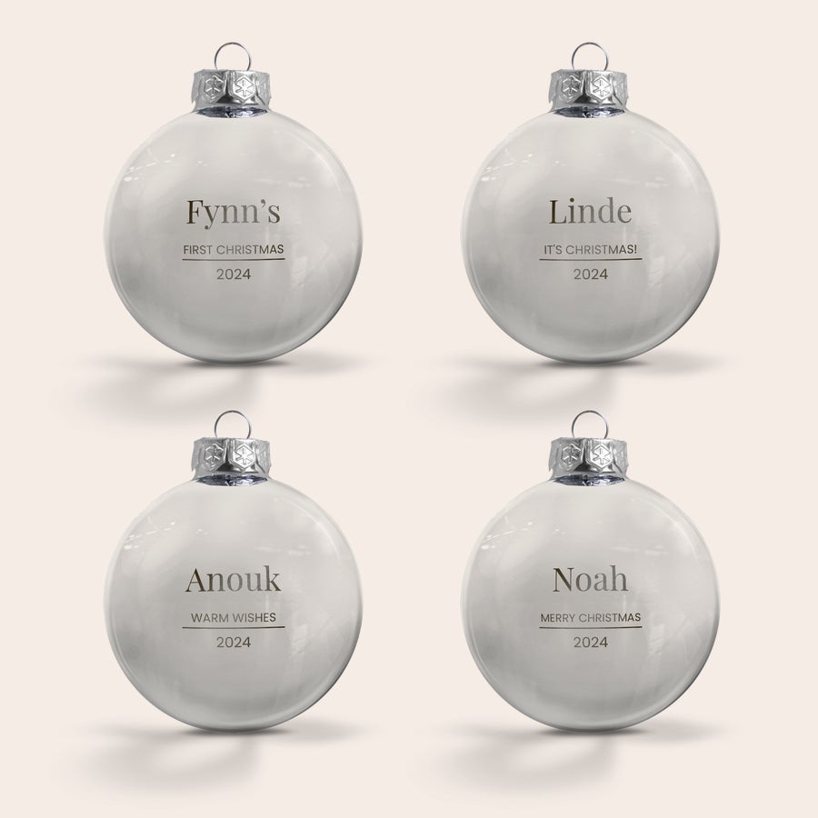 Glasjulkulor med namn Fyra graverade julgranskulor i glas med silverfärgade fattningar och personliga namn och datum, som "Fynn's First Christmas 2024" och "Linde It's Christmas! 2024". Ge din julgran en personlig touch med en graverad julgranskula.