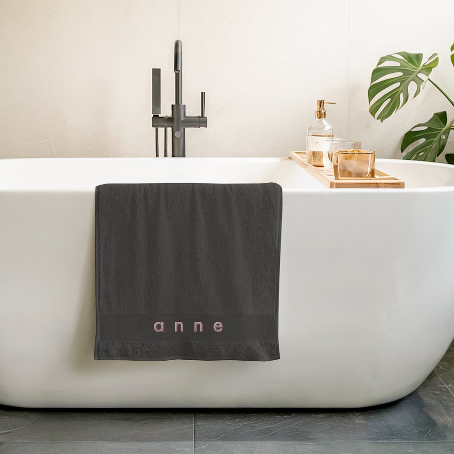 Serviette brodée Serviette de bain gris anthracite brodée avec le prénom Anne en rose pâle, suspendue sur une baignoire blanche