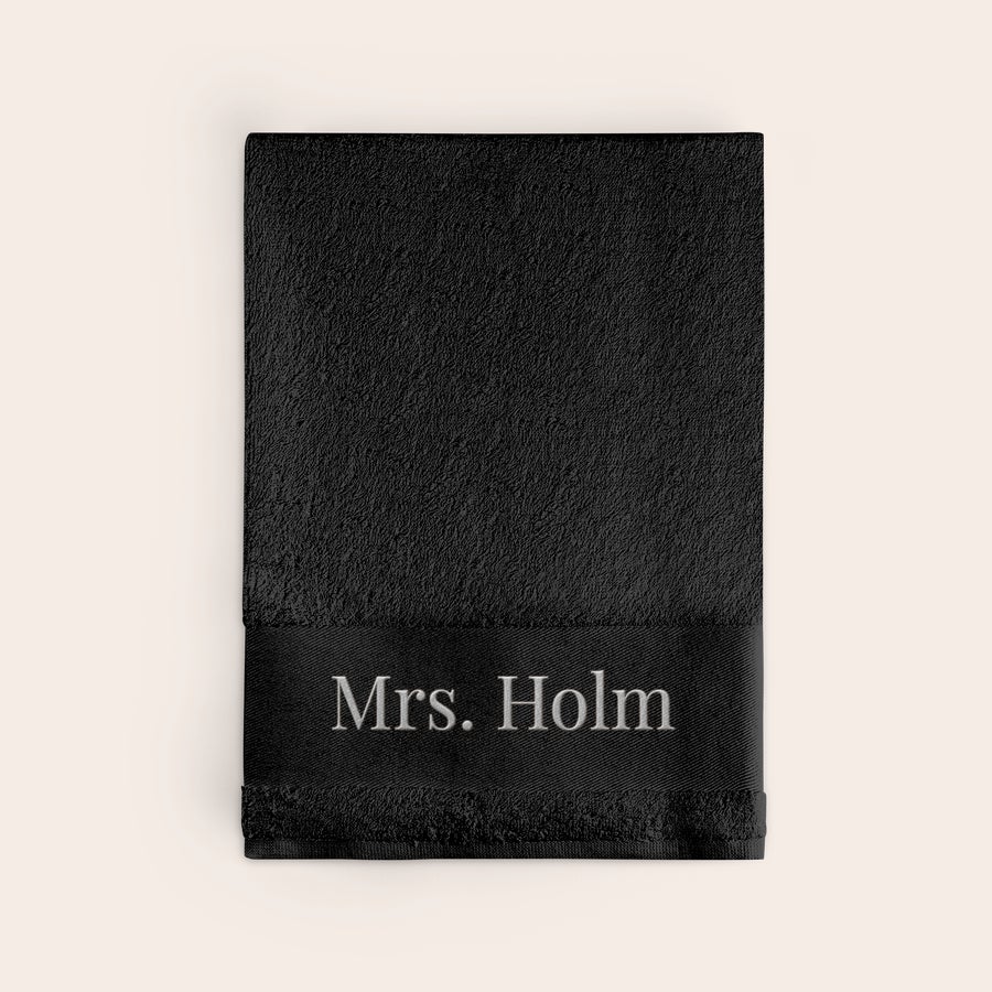 Badehåndklæde med tekst Sort personligt badehåndklæde med teksten "Mrs. Holm" broderet i hvid skrift langs kanten.
