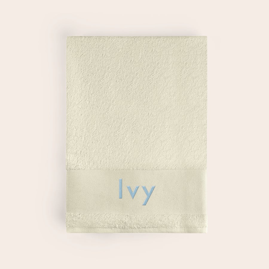 Bath towel with text Folded beige bath towel embroidered with the name Ivy in light blue, a personalized beige bath towel