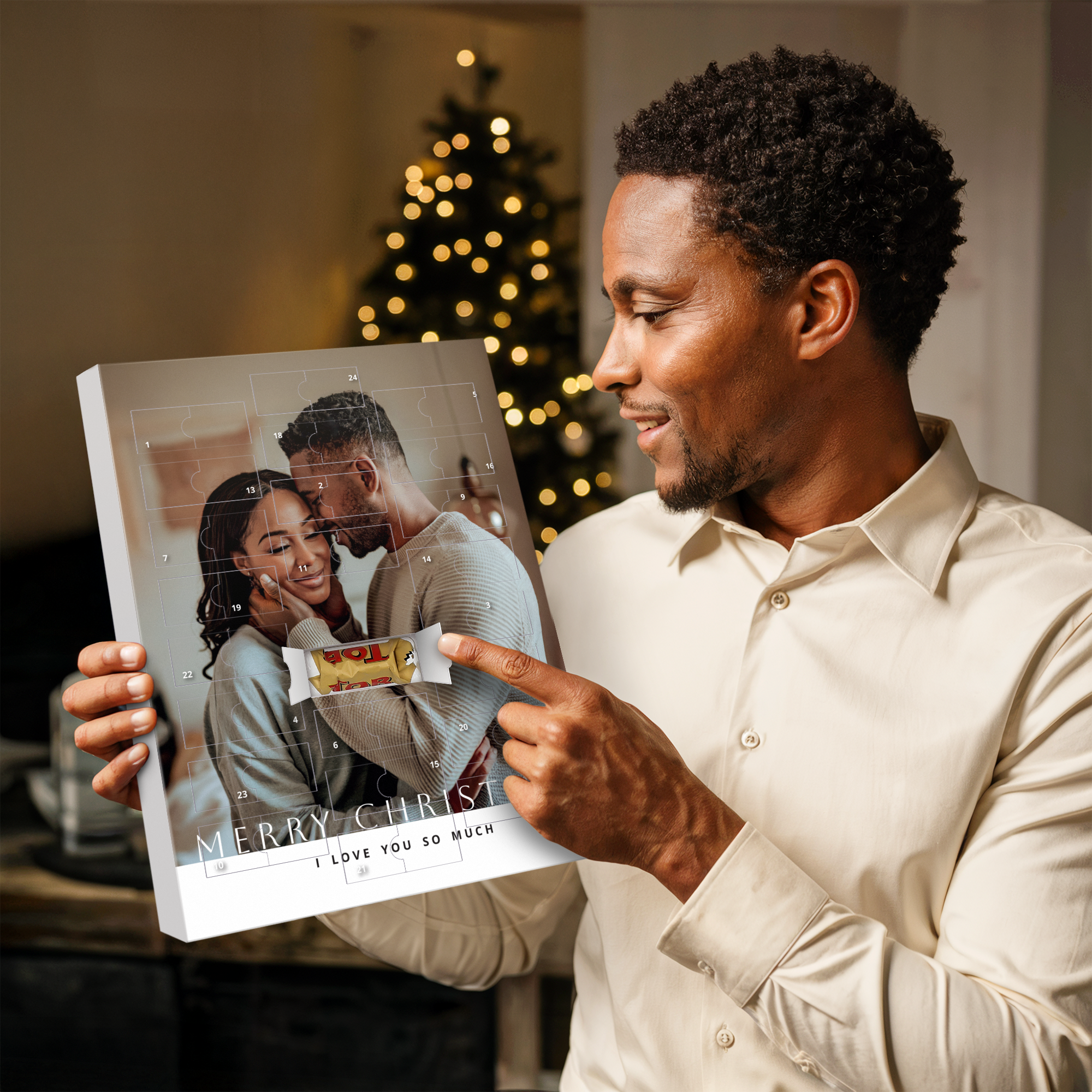 A man holding a personalized advent calendar with a printed photo of a couple and the text "MERRY CHRIST I LOVE YOU SO MUCH" pointing to an opened window with a Toblerone chocolate bar, counting down to Christmas.
