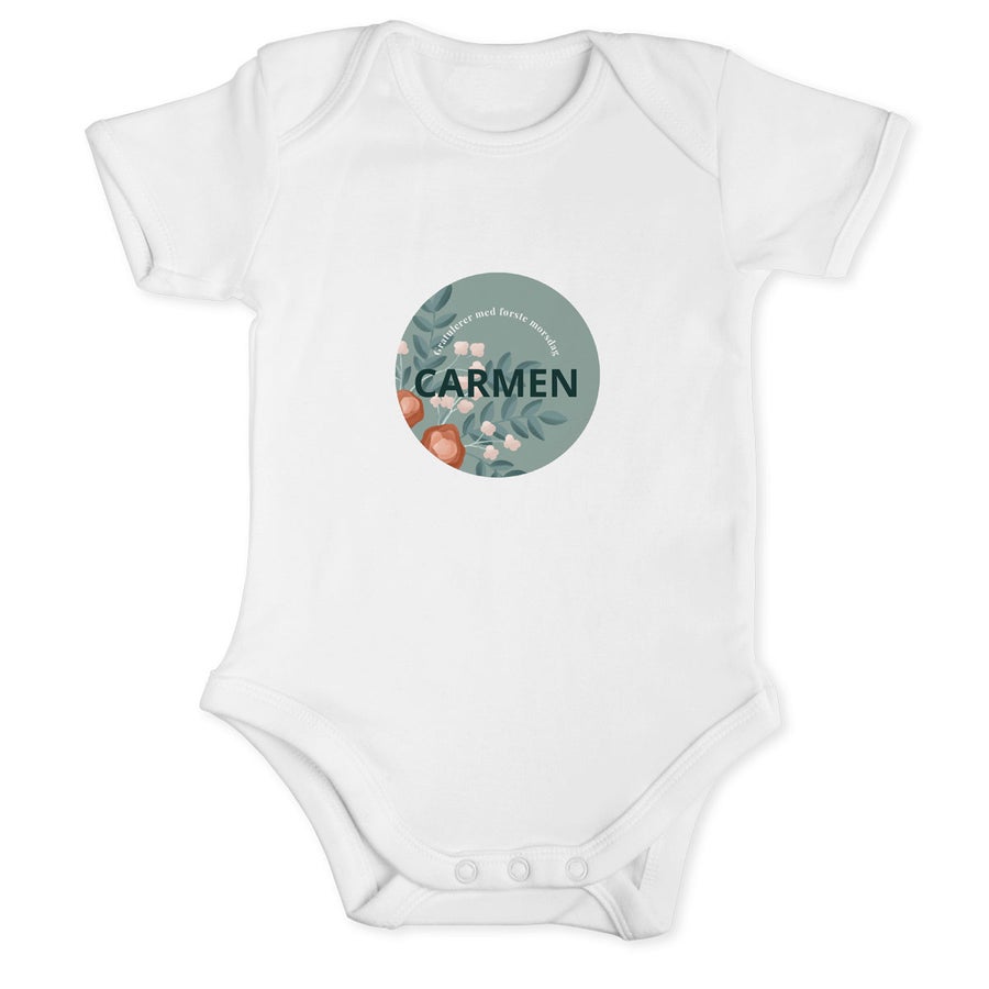 Hvit personlig trykt onesie med navnet CARMEN og Gratulerer med første morsdag, omkranset av et blomstermønster.