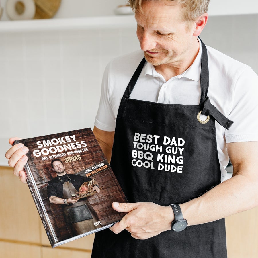 Smokey Goodness BBQ-Buch Mann mit Schürze die “Best Dad Tough Guy BBQ King Cool Dude” bedruckt ist hält ein personalisiertes Smokey Goodness Grillbuch für Jonas in den Händen