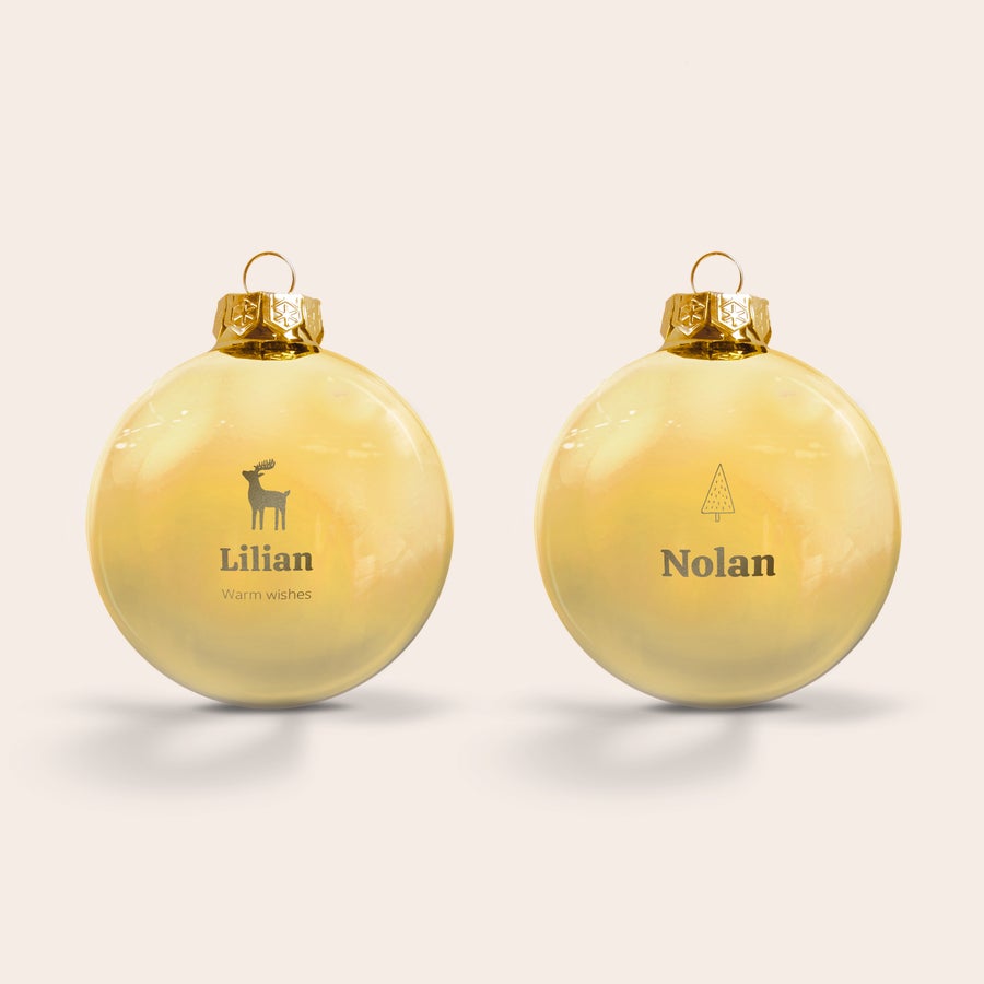 Vianočné gule - sklenené Dve zlaté sklenené vianočné gule, jedna s rytým sobom a menom Lilian s textom Warm wishes, druhá s vianočným stromčekom a menom Nolan, ideálne na ozdobenie vianočného stromčeka prispôsobenou vianočnou cetkou.
