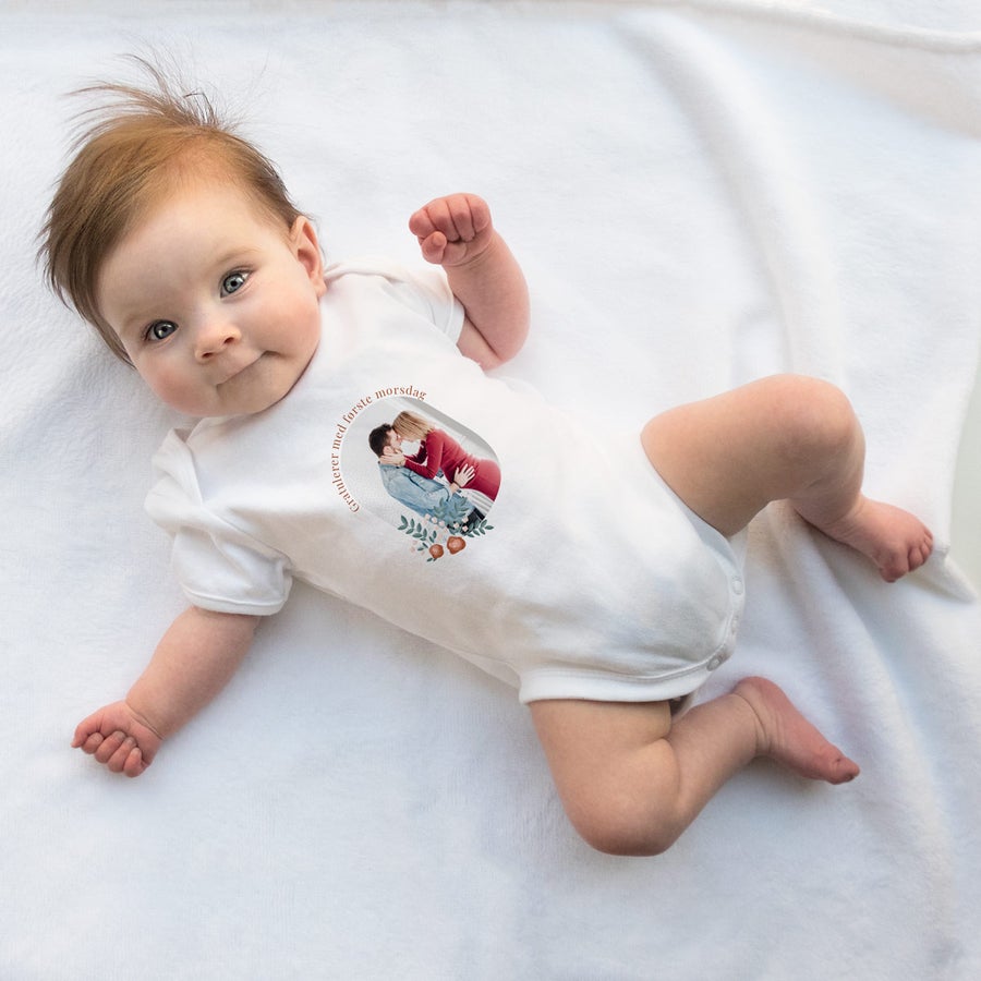 Baby i hvit personlig trykt onesie med bilde og gratulerer med første morsdag-tekst.