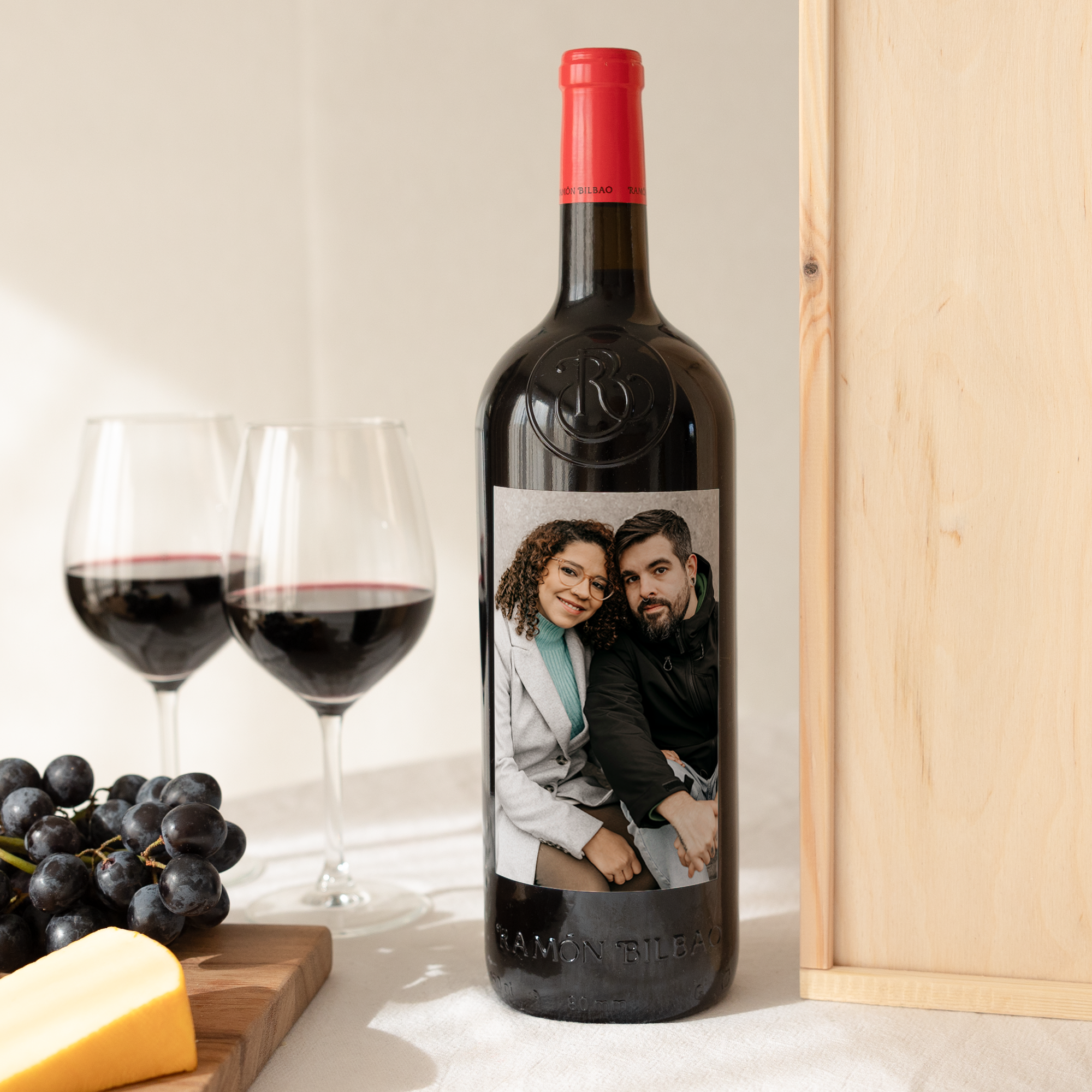 Sticlă de vin Ramon Bilbao cu etichetă personalizată printată cu fotografia unui cuplu, lângă pahare de vin și struguri.
