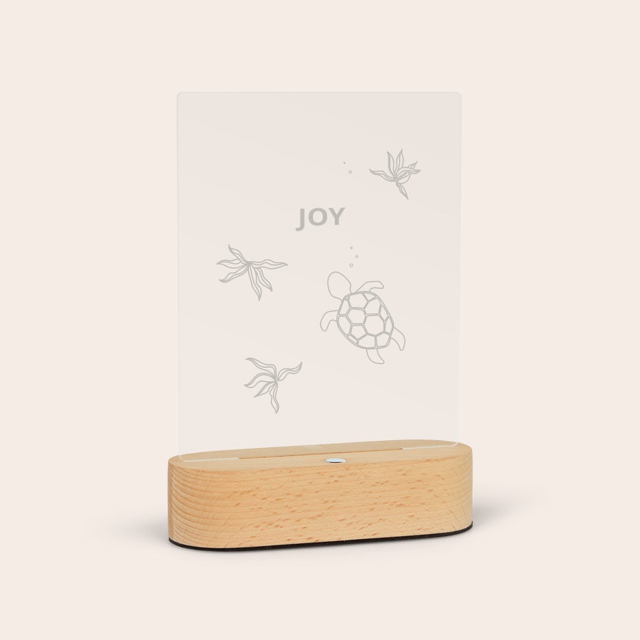 Lamp met naam Transparante gegraveerde lamp met een houten standaard, versierd met een schildpad en zeewier. Met het woord 'JOY' bovenaan de lamp met gravure.