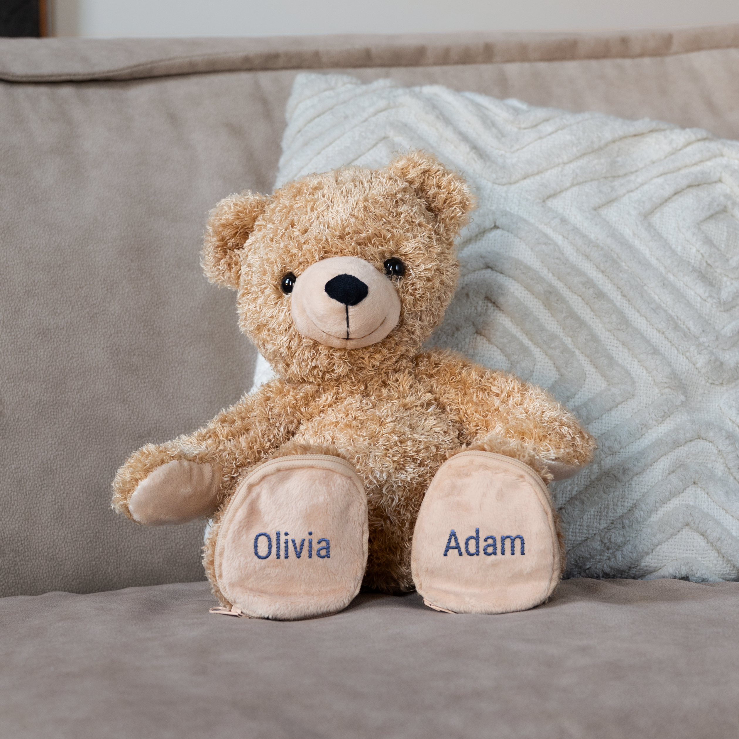 Brauner Teddybär mit Namen Olivia und Adam auf den Füßen gestickt, sitzend auf einem Sofa