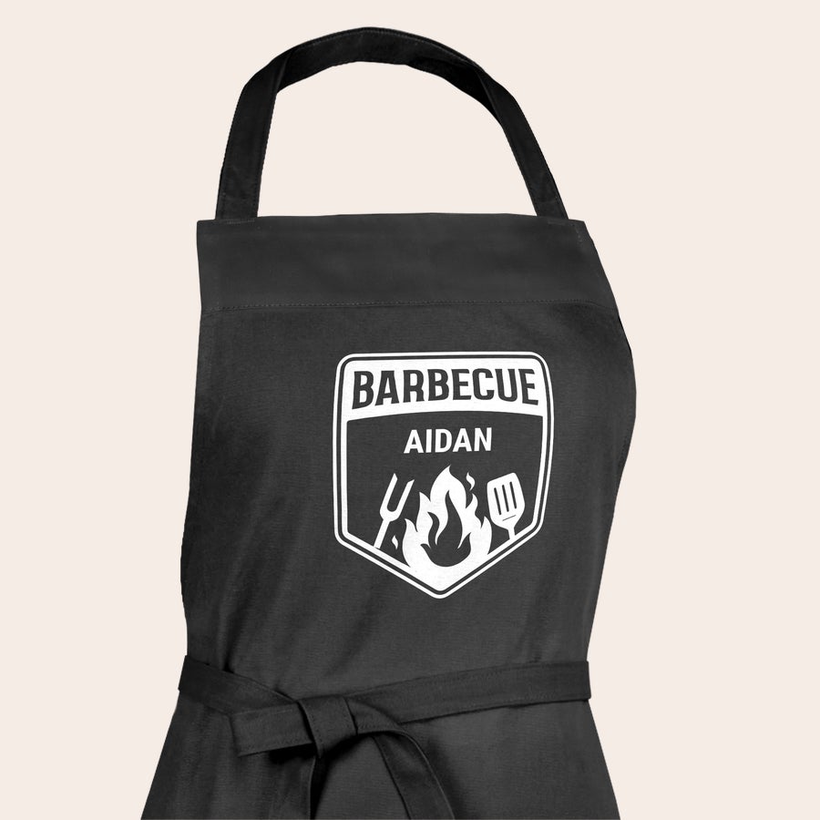 Șorț pentru grătar personalizat - negru Șorț de grătar personalizat negru, cu textul "BARBECUE AIDAN" imprimat alb și flăcări. Proiectează-ți propriul șorț.