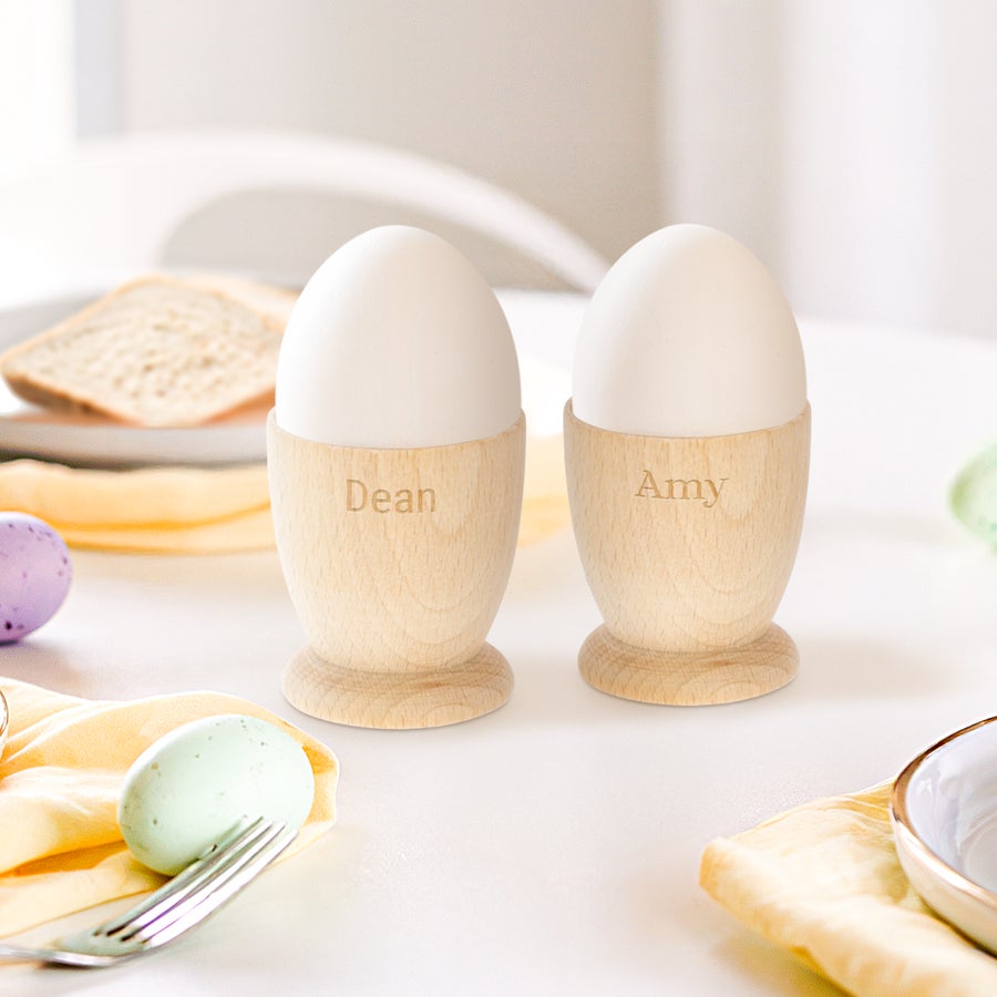 Huevera personalizada Dos hueveras personalizadas de madera de haya con los nombres Dean y Amy grabados, cada una con un huevo blanco.