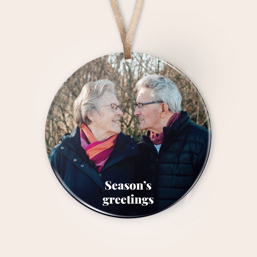 Személyre szabott plexi karácsonyi díszek Személyre szabott plexi karácsonyi dísz egy idős pár fotójával és "Season's greetings" felirattal, szalaggal felakasztva.