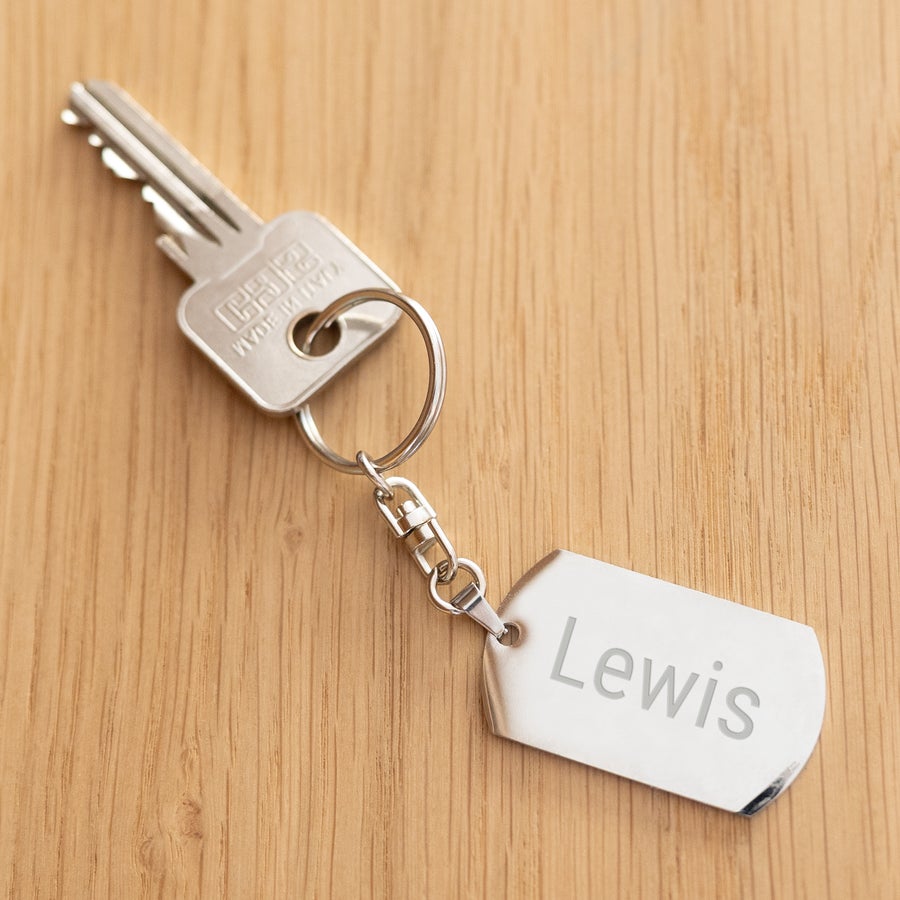 Llavero grabado Llavero personalizado con grabado, muestra el nombre "Lewis" grabado en un llavero metálico rectangular con un llavero redondo y una llave, todo sobre una superficie de madera clara.