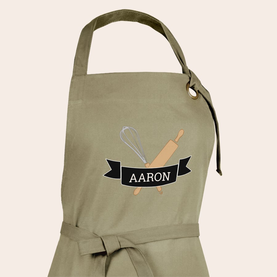 Tablier personnalisé Tablier personnalisé beige avec le prénom AARON imprimé sur un ruban noir, un fouet et un rouleau à pâtisserie.