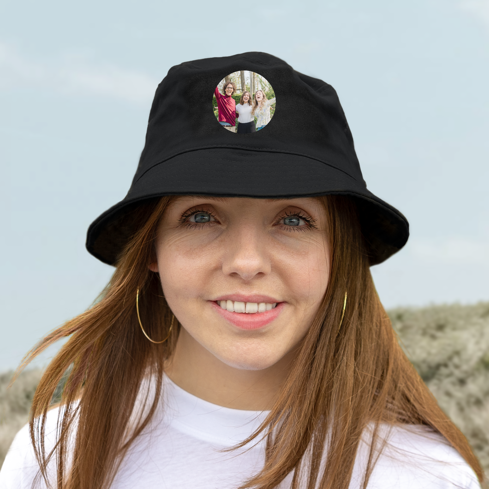 Vrouw met bruin haar en gouden oorbellen draagt een zwarte bucket hat bedrukt met een foto van drie vrienden, voor een unieke look.