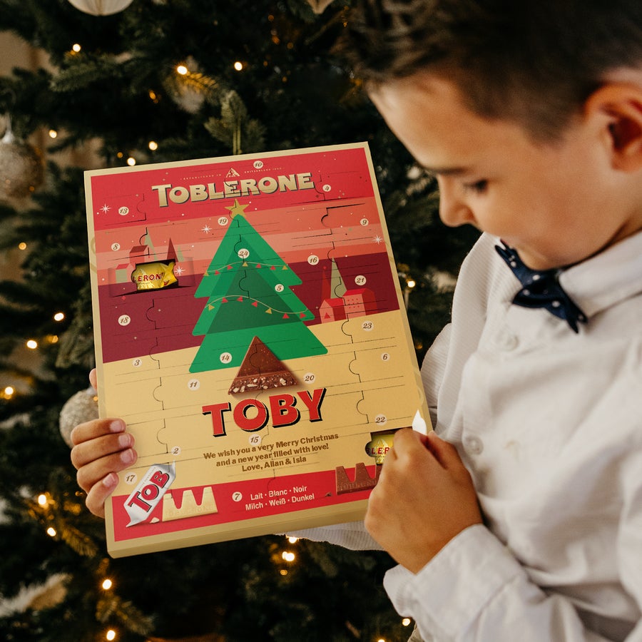 Calendrier de l'avent Toblerone  2025 Enfant ouvrant un calendrier de l'Avent Toblerone personnalisé avec le prénom Toby et un message sous un sapin de Noël illuminé.