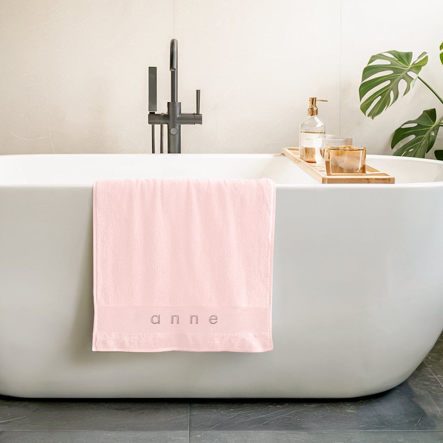 Handduk med Text En rosa handduk med broderat namnet Anne i grått, hänger över kanten på ett vitt badkar. Gör varje badstund extra personlig med en rosa handduk som är broderad.