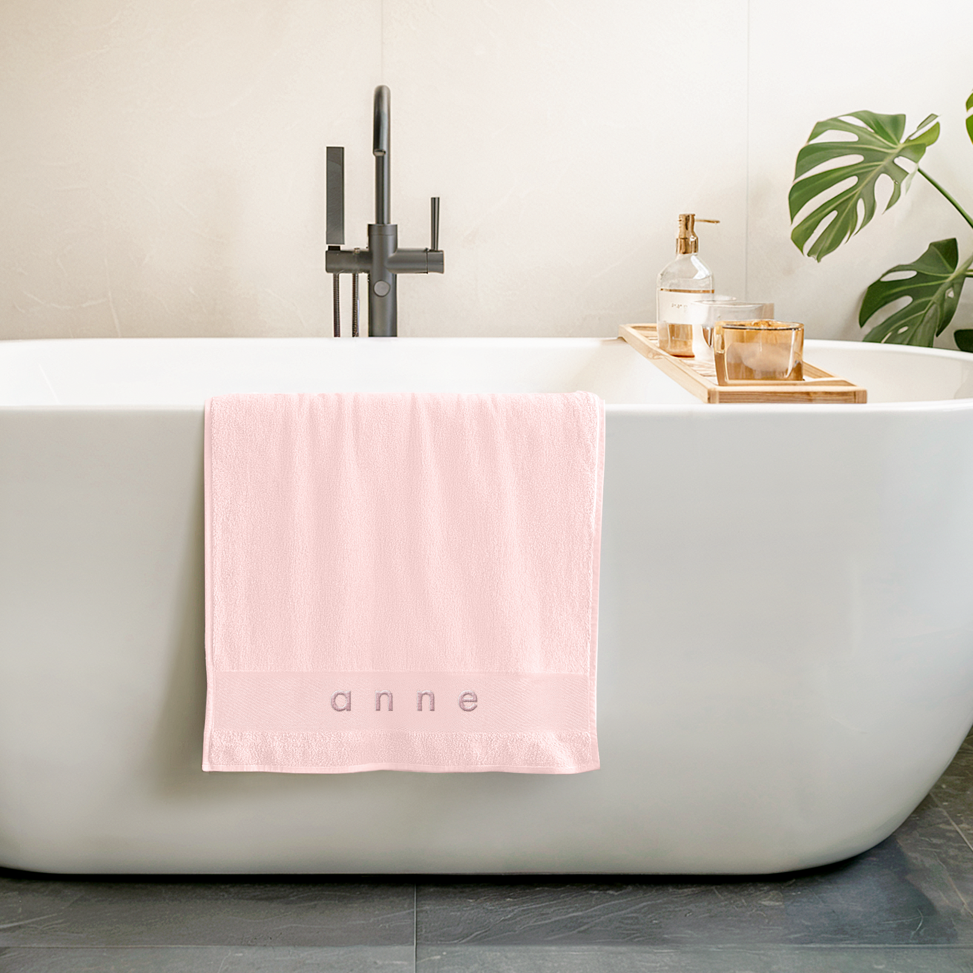 Toalla rosa claro personalizada con el nombre Anne bordado en gris, colgando de una bañera blanca.