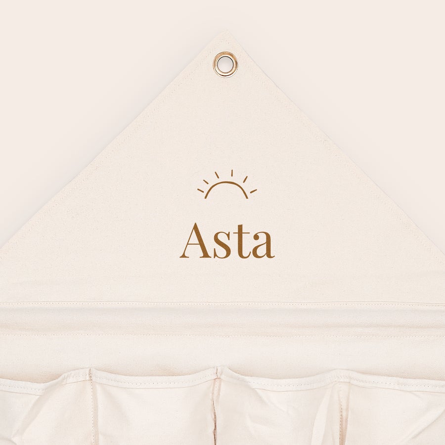 Hængende opbevaring med navn og/eller billede Personaliseret hængende opbevaring i offwhite stof med "Asta" og sollogo printet i brun skrift. Få styr på alle dine ting.