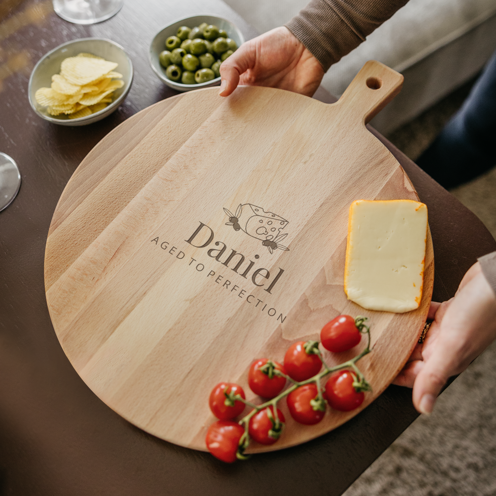 Tabla de servir de madera redonda con grabado láser del nombre Daniel y queso, aceitunas, tomates y patatas fritas.