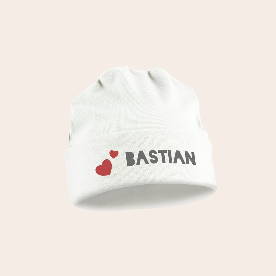 Babymütze mit Namen Weiße Babymütze bedruckt mit dem Namen BASTIAN in Großbuchstaben und zwei roten Herzen als süßes Design.