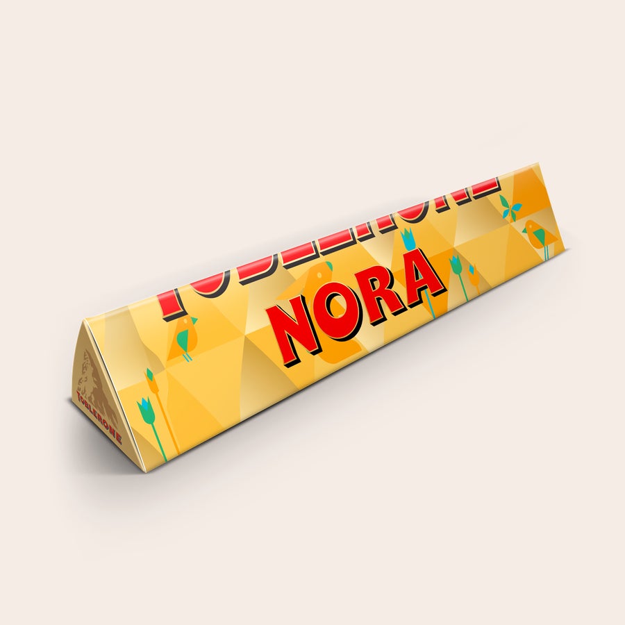 Toblerone med navn Personlig Toblerone chokolade med "NORA" trykt på indpakningen i rødt med guldskygge.